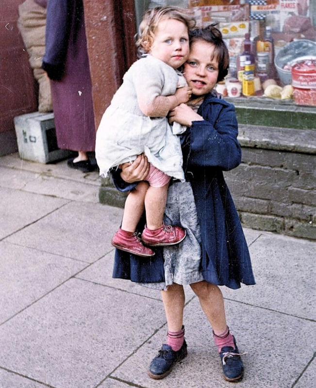 [colorized] Taken on the 20th February, in 1954. for Picture Post magazine, city street photographs . 
A little girl pictured with her younger sister on a street in Belfast.